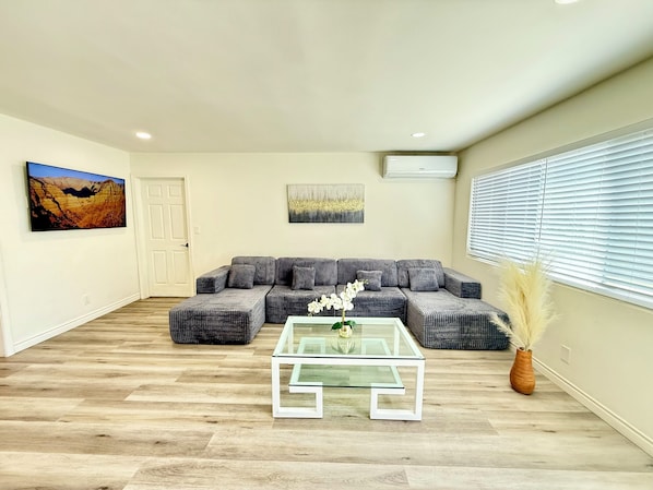 A living room with a sectional sofa, a glass coffee table, a painting, and a window with blinds.