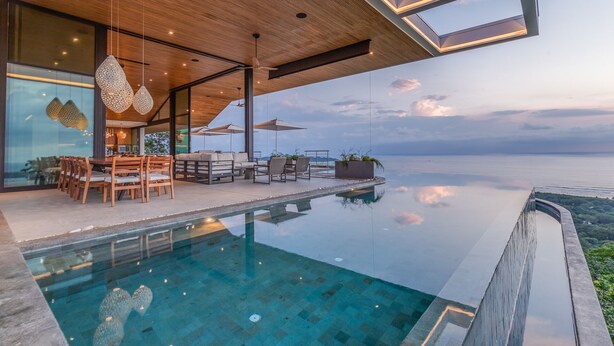 Zona de piscina exterior moderna con techo de madera, asientos y vistas al mar.