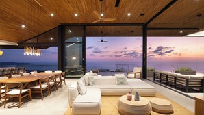 Un interior moderno con techo de madera, un sofá grande, una mesa de comedor y vistas al océano al atardecer.
