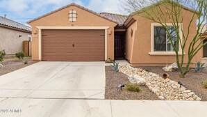 Une maison de plain-pied avec une porte de garage marron, un petit jardin devant la maison agrémenté de rochers et d’un cactus, et un ciel dégagé.