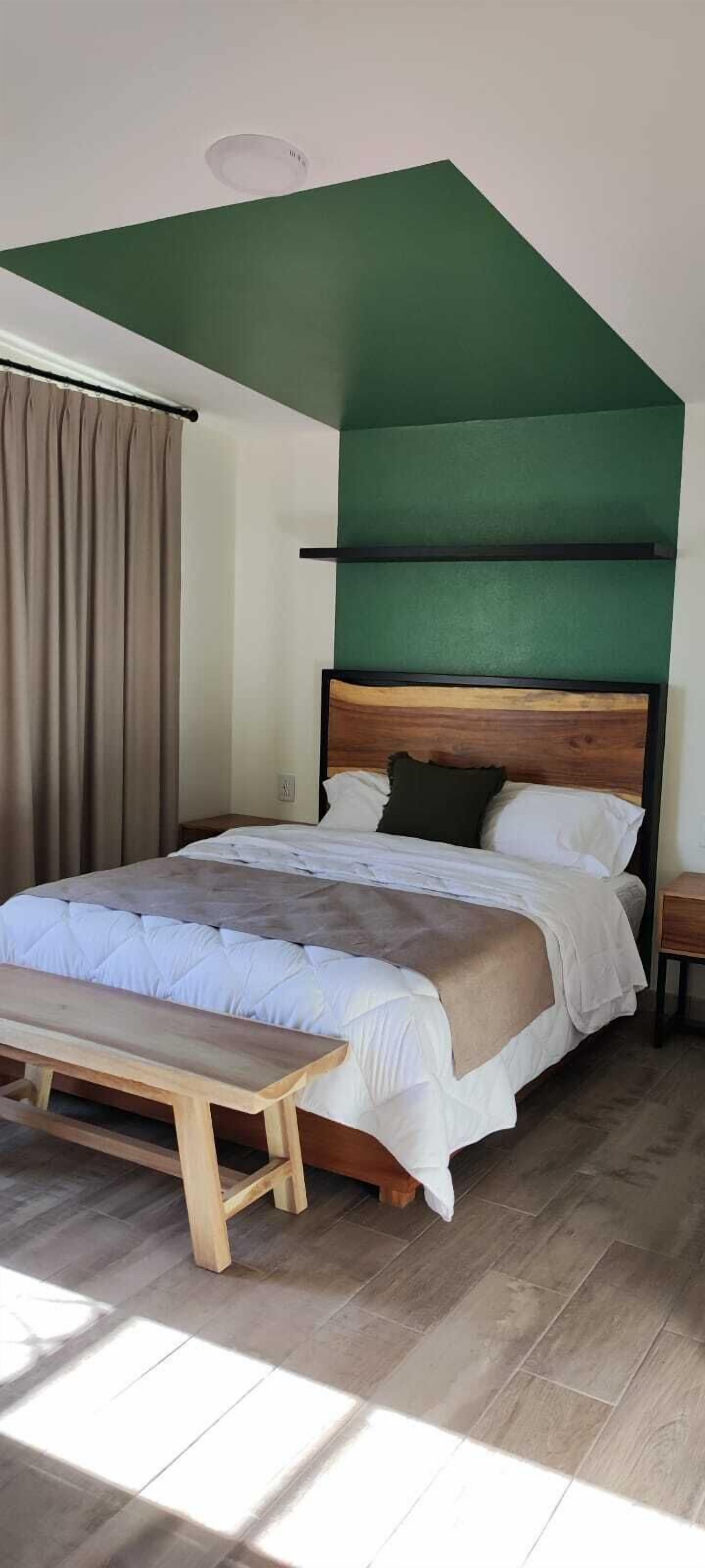 Quarto com cama, um banquinho, uma mesa de cabeceira e uma parede verde.
