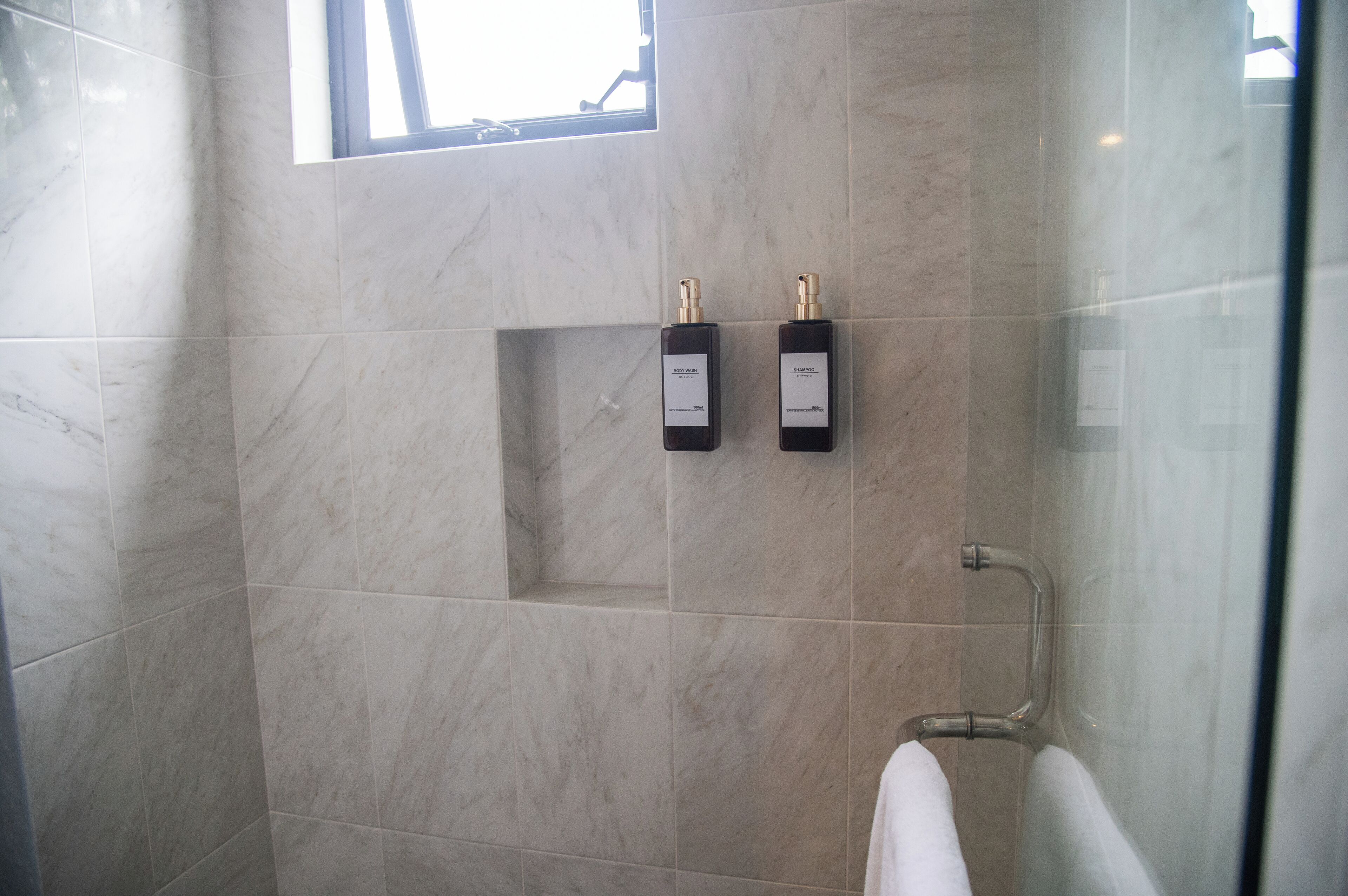A bathroom with marble tiles, a glass shower enclosure, and two bottles of shampoo on the wall.