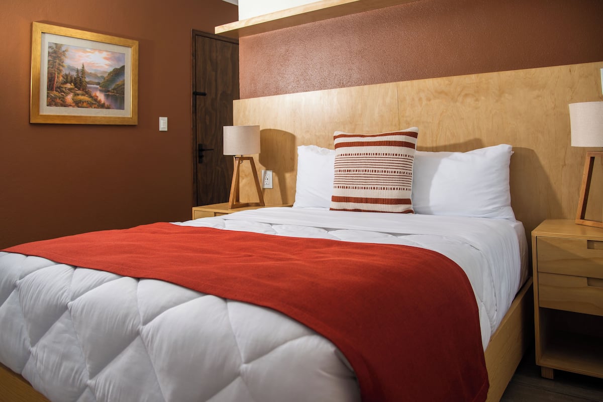 A neatly made bed with a red blanket, white pillows, and a wooden headboard. There is a framed picture on the wall and a bedside table with a lamp.
