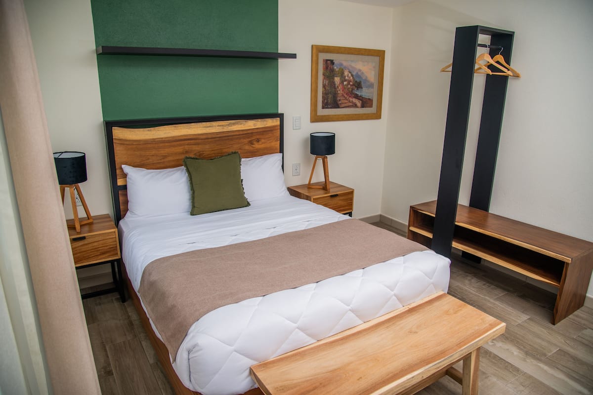 A neatly made bed with a wooden headboard, a bedside table with a lamp, a green pillow, and a painting on the wall.