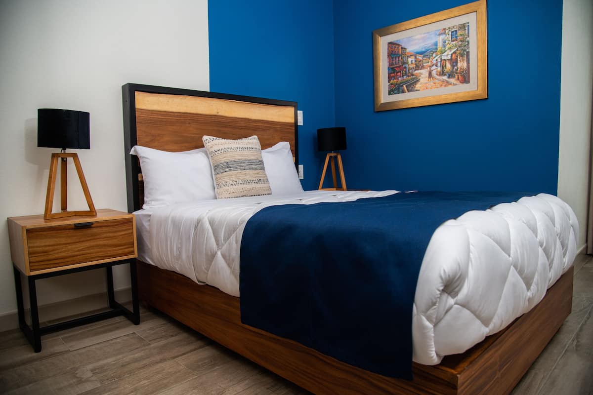 A neatly made bed with a blue blanket and white pillows, flanked by two bedside tables with lamps, in a room with blue walls and a framed picture above the bed.