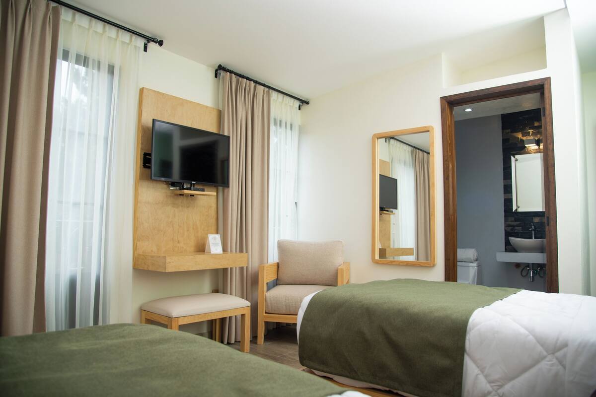 A hotel room with two beds, a television, a chair, a small table, and a bathroom visible through an open door.