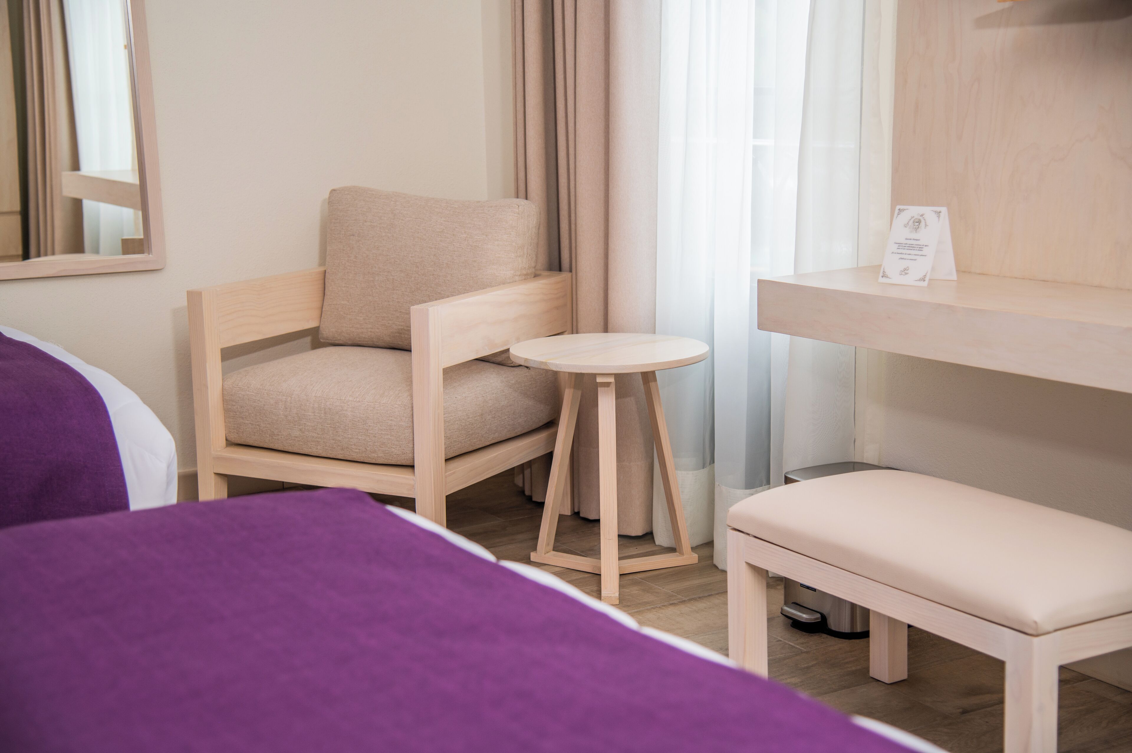 Quarto de hotel com cama, cadeira, mesa pequena e um banquinho.