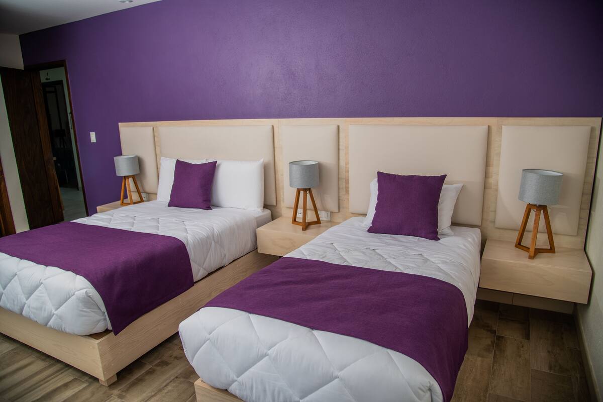 A hotel room with two beds, each with a wooden headboard, white bedding, and purple accents. There are bedside lamps on wooden stands.