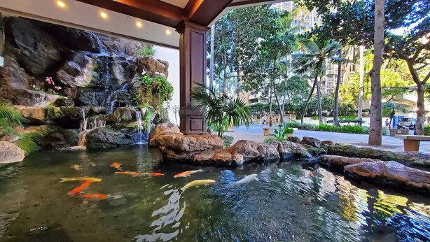 A koi pond with a waterfall and rocks, surrounded by greenery and a building in the background.