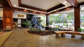 A lobby with a stone floor, a wooden bench, a waterfall feature, and a sign for 'Tower 1 Makai'.