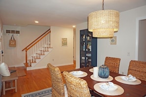 A dining area with a wooden table, wicker chairs, and a hanging light fixture. - Wedgewood Cottage On the Cape (Barnstable)