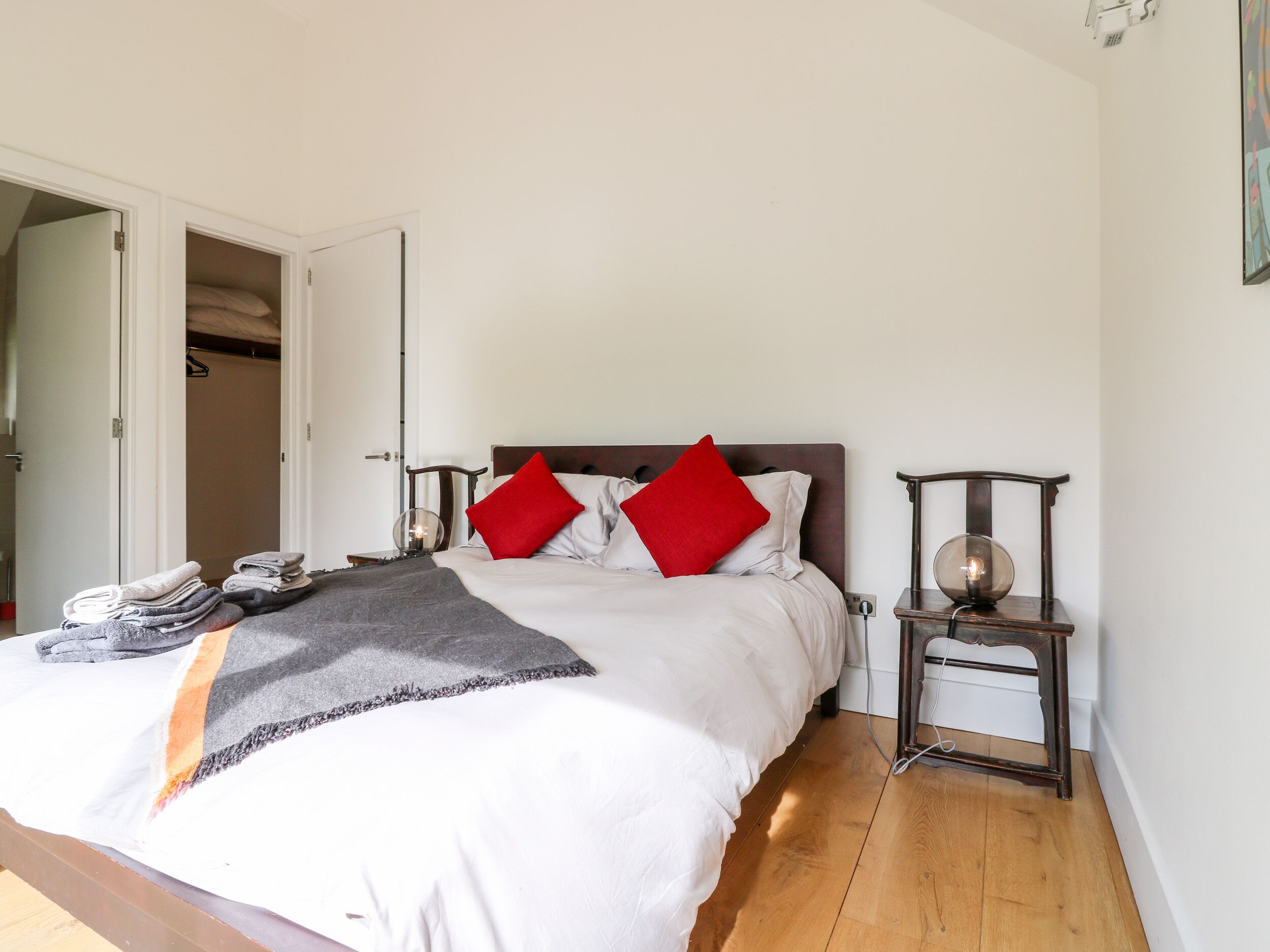 A neatly made bed with a grey blanket and two red pillows, a bedside table with a lamp, and a wooden floor.