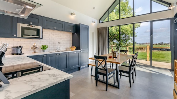 A modern kitchen with dark cabinets, a marble countertop, and a large window overlooking a green landscape.