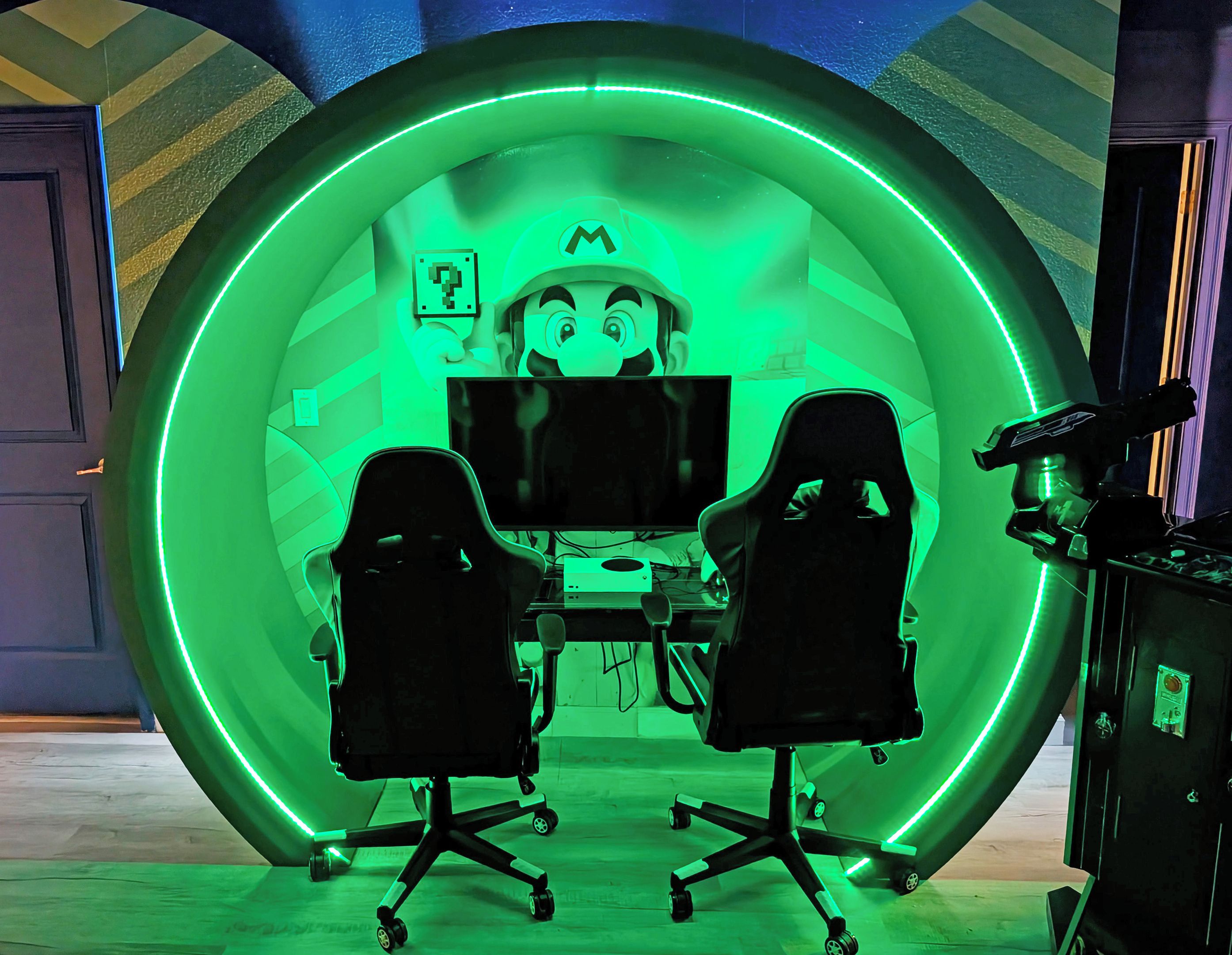 A room with a circular green-lit structure, two gaming chairs, a desk with a computer, and a gaming console.