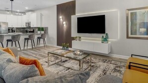Una sala moderna con sofá, mesa de centro y televisor montado en la pared. Se observa una cocina al fondo con mesa y sillas.