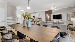Una sala moderna con una gran mesa de comedor de madera, sillas tapizadas de gris, un sofá gris y un televisor de pantalla plana montado en la pared.