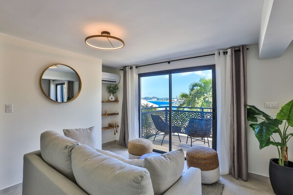 A modern living room with a sofa, round mirror, and a view of a balcony with outdoor furniture.