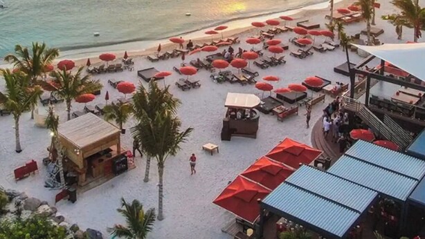 Un complexe hôtelier situé en bord de mer, doté de chaises longues et de parasols, d’un bâtiment en bois et offrant une vue sur la mer et les bateaux.