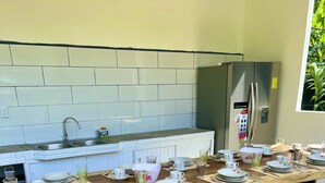 A long wooden dining table set with plates, cups, and cutlery, in a kitchen with a white tiled backsplash and a stainless steel refrigerator.