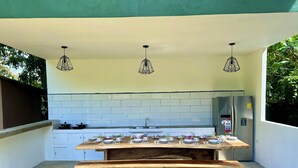 An outdoor kitchen with a dining table set for four, a stainless steel refrigerator, and hanging pendant lights.