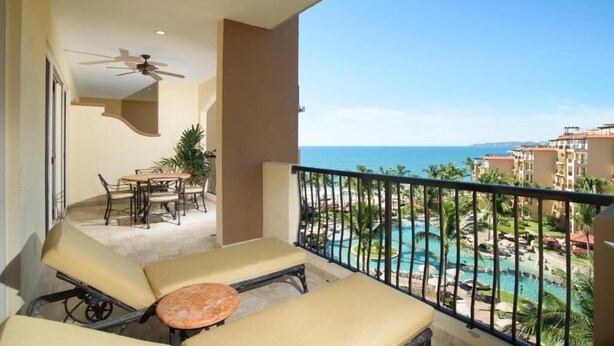 Un balcone con poltrone relax e un tavolo, affacciato su una piscina e gli edifici del resort.