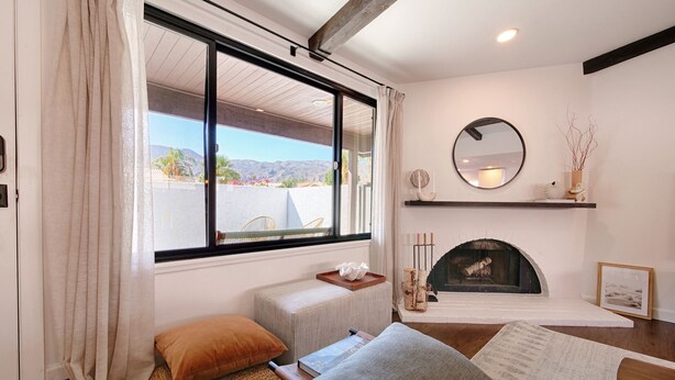 A cozy living room with a large window, a fireplace, a comfortable sofa, and a wooden floor.
