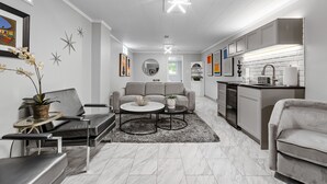 Un salon moderne avec un canapé, une table basse et des fauteuils, une cuisine équipée de meubles et d’appareils électroménagers, et un couloir avec une porte donnant sur une autre pièce.