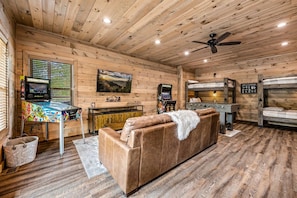 A cabin interior with a wooden ceiling, a sofa, a table with a colorful tablecloth, a television, and bunk beds.