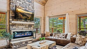 A cozy cabin interior with a stone fireplace, leather sofa, and large windows offering a view of trees. - Top 5% · Pet Friendly · Hot Tub · Arcade · Views (Sevierville)