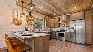A modern kitchen with wooden cabinets, a stainless steel refrigerator, and a bar area with stools. - Top 5% · Pet Friendly · Hot Tub · Arcade · Views (Sevierville)