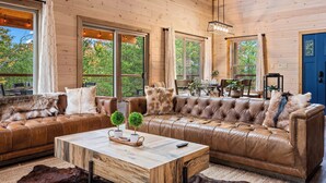 A cozy cabin interior with a large leather sofa, a wooden coffee table, and large windows offering a view of the outdoors. - Top 5% · Pet Friendly · Hot Tub · Arcade · Views (Sevierville)