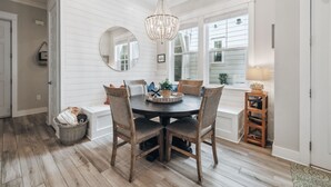 Un coin repas comprenant une table ronde, des chaises, un lustre et une grande fenêtre avec des stores.
