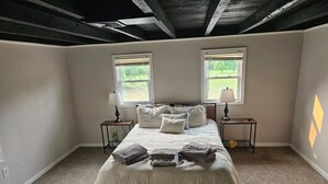 Un dormitorio con una cama grande, dos mesitas de noche, dos ventanas y un techo con vigas a la vista.