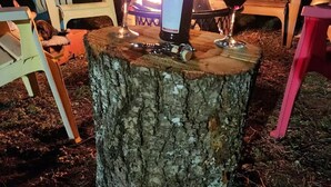 Un entorno rústico al aire libre con un brasero rodeado de sillas de madera y una botella de vino.