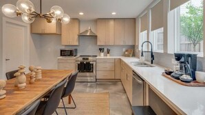 A modern kitchen with a wooden dining table, stainless steel appliances, and a large window providing natural light. - Rooftop Fire Pit + Bar ✨ Near Broadway (Nash)
