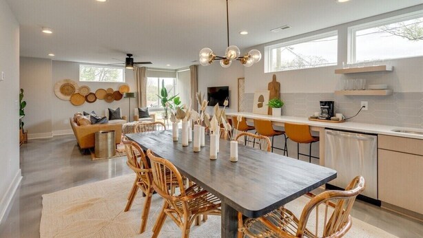 A modern kitchen with a dining area, featuring a long wooden table, wicker chairs, and a central island with a sink. - Rooftop Fire Pit ~ Bach Parties ~ Sleeps 24! (Nashville)