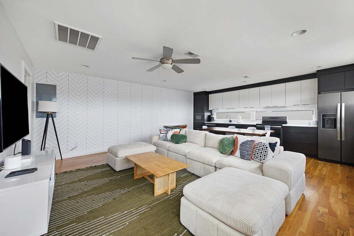 Ein modernes Wohnzimmer mit einer Couch, einem Hocker und einem Couchtisch, einem Fernseher und einer Küchenzeile mit Schränken und Geräten.