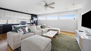 A modern living room with a sofa, ottoman, and coffee table. There is a kitchen area with cabinets and a refrigerator. A ceiling fan is visible, and there are windows on the wall.