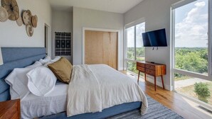 Ein Schlafzimmer mit einem Bett, einem Fernseher und einem großen Fenster mit Blick auf Bäume.