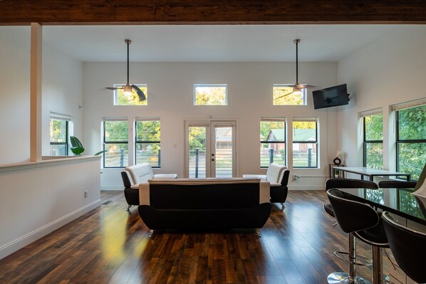 Una habitación amplia con un sofá negro grande, una mesa de comedor y varios ventanales que dejan entrar luz natural.