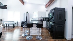Eine moderne Küche mit einem schwarzen Kühlschrank, einem Doppelbackofen und einer Kaffeemaschine. Zwei schwarze Barhocker und ein weißer Esstisch mit Stühlen stehen bereit. Die Küche verfügt über weiße Schränke und eine Holzbalkendecke.