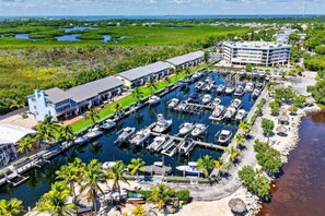 A marina with multiple boats docked, a building with a flat roof, and a body of water with greenery in the background.