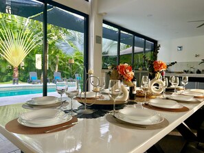 A dining table set with white plates, gold cutlery, and wine glasses, overlooking a pool and lush greenery.