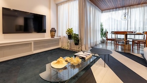 A modern living room with a flat-screen TV, a glass coffee table with decorative items, a dining area with wooden furniture, and large windows with sheer curtains. - 10 Minutes From Ipanema Beach Vm201 (Rio de Janeiro)