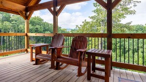 Eine Holzterrasse mit zwei Adirondack-Stühlen und einem kleinen Tisch, mit Blick auf einen bewaldeten Bereich.