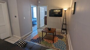 A living room with a sofa, a coffee table, a TV, and a floor lamp. There is a door leading to another room. - 2BR Apt Right on the Town Square (Robinson)