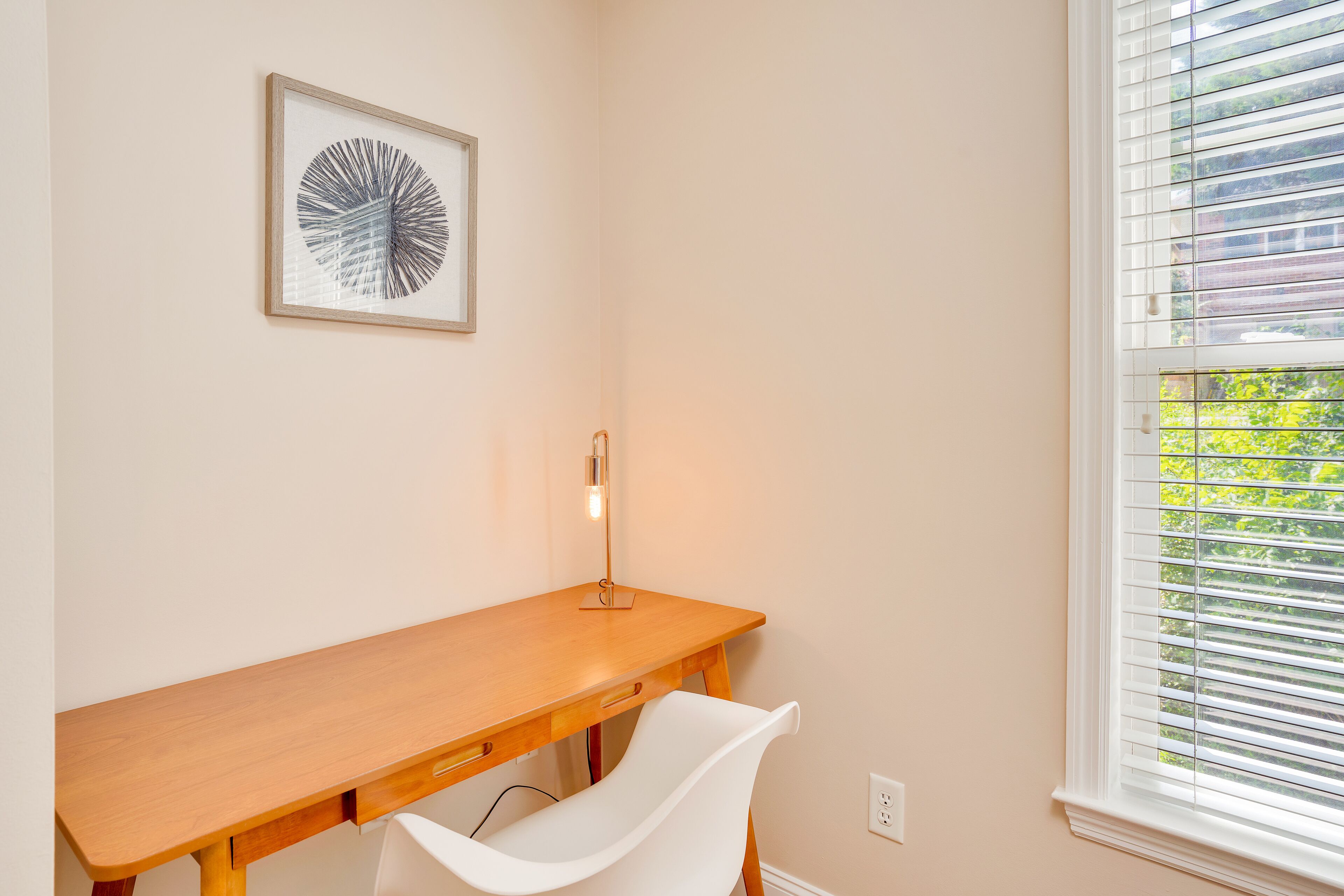 A corner room with a wooden desk, a white chair, a framed artwork, and a window with blinds.