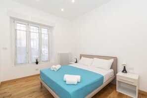 Moderni hotellihuone, jossa on suuri sänky, yöpöytä, ikkuna, jossa on sälekaihtimet, ja patteri.