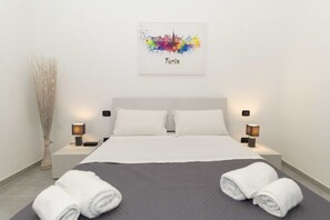 Habitación de hotel con cama, dos mesitas de noche, una obra de arte con vista urbana en la pared y un jarrón decorativo.
