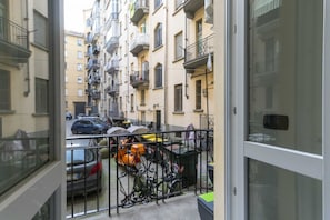 A narrow urban street with parked cars and bicycles, multi-story buildings with balconies, and a metal fence.